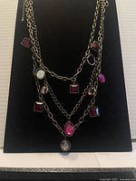 Front view of the gold-tone multi-layer chain necklace with red and oval gemstone charms displayed on a black stand.