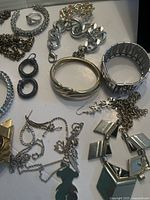 Photo of assorted silver and mixed metal jewelry including bracelets, rings, necklaces, and earrings spread on white surface.