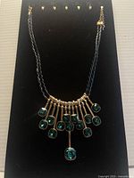 Front view of brass necklace featuring blue gem-like stones and black braided chain hanging vertically on black background.