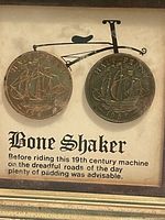 Close-up photo of the two vintage half penny coins mounted as bicycle wheels with the 'Bone Shaker' text and illustration.