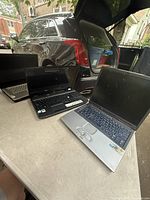 Three laptops positioned outdoors near a car trunk with all screens visible except one closed laptop.