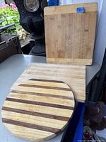 Three solid wood cutting boards displayed, showing varying shapes: square, rectangular, and round with stripes.