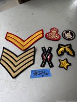 Seven vintage embroidered military patches laid out on a grey surface, including chevrons, crossed rifles, star, and crests.