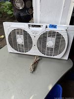 Front view of white double window fan with two fan grills and power cord placed on a gray surface.