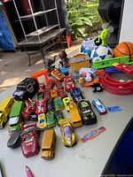 Overview of multiple die cast cars and various kids toys arranged on a table outdoors, showing variety of vehicles and additional plastic toys.
