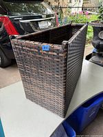 Photo of the outside front corner and side of the dark brown woven rattan box sitting on a table outdoors near a car.