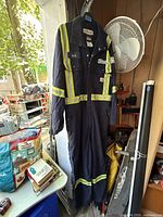 Front view of the navy blue Carhartt coverall with reflective yellow stripes hanging on a black hanger next to a fan and various items.