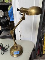Solid brass adjustable desk or bedside lamp shown from side view on a table, illustrating the overall shape, adjustable arm joints, round weighted base, and dome shade.