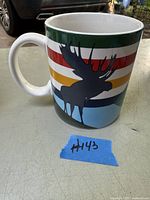 View of the coffee cup showing the multicolor horizontal stripes and black moose silhouette on white ceramic surface.