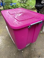 A large pink plastic storage tote with a rectangular lid, situated outdoors on concrete with some green foliage in the background. The tote appears in good condition with visible water droplets on the lid.
