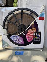 Front view of coiled LED light strip on black reel in damaged retail box with an image of the multi-color LED lights and remote control on the box front.