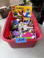 A red plastic bin filled with assorted LEGO Classic bricks, plates, and pieces in various colors, with an instruction booklet for set 10704 placed inside the bin.