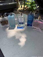 Six assorted men's cologne and perfume bottles on a white table outdoors, various shapes and colors including blue and pink bottles.