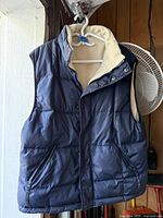 Front view of navy blue men's winter vest with beige fleece lining on hanger, showing five pockets and snap button closure