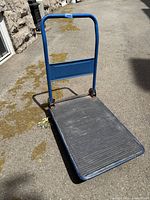 Blue metal folding moving cart in open position placed on pavement showing rubberized platform and handle.