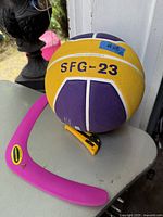 Side view of yellow and purple Nike basketball labeled SFG-23 with a crown symbol and pink Aerobie boomerang on a gray surface.