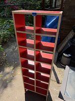 Photo of a tall, narrow wooden storage cupboard with red shelves and no back panel, placed outdoors.