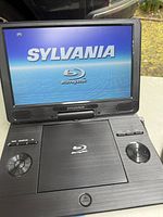Front view of the open Sylvania portable Blu-ray player displaying the Sylvania start-up screen on the built-in LCD.