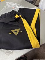 Close-up of folded black sweatpants with yellow side stripe and triangular emblem with arrows on front.