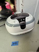 White and gray ultrasonic jewelry cleaner with lid closed, visible on/off button and power cord attached, placed on a gray surface.