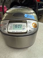 Front view of Zojirushi stainless steel rice cooker showing digital display with time 16:03 and control buttons.