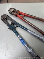 Two vintage bolt cutters placed on a gray surface, one with red handles and one with dark blue handles wrapped in tape, showing signs of wear.