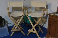 Photo showing two wooden directors chairs with beige canvas seats and backs placed next to each other on blue carpet.