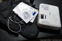 Epson LCD projector placed on black surface with power and AV cords, manuals, and carrying bag.