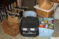Photo showing various baskets including wicker and woven plastic bins, a black insulated bag, and two folding luggage stands stacked behind, a blue and white woven plastic bin filled with colorful packing tape rolls