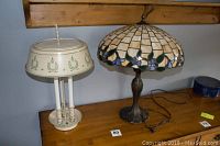 Photo of two table lamps on a wooden surface against a gray wall, showing the Tiffany style stained glass lamp and the metal tole painted lamp side by side.