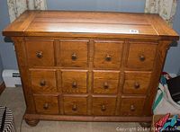 Front view showing 12 small square drawers with round wooden knobs, wooden panel top and carved details on corners and base.