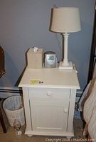 Front view of one white end table displaying one drawer and paneled cabinet door with round knobs; lamp with white textured shade on top.