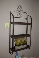 Brown metal wall shelf mounted on wall with decorative scroll top and two shelves containing a wheat display and a blue bottle