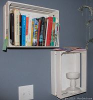 Two whitewashed wooden crates mounted on a wall, one holding books and the other holding a white pedestal bowl.