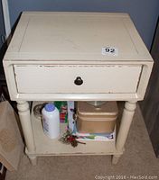 Photo of one cream-colored country style end table showing square top, single drawer with round knob, turned legs, and lower shelf with items stored on it. Visible paint chipping and wear.