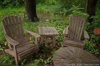 Shows overall set of two Adirondack chairs and square side table outdoors surrounded by grass and trees, wood weathered and chipped.