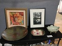 Two framed pictures: multi-colored 3D paper flower with gold frame, black-and-white outdoor photo print in black frame, along with three decorative dishes placed on a black table.