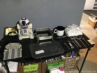 Image of black tabletop showing 4 purses, R2D2 cookie jar, silver toned pan holder, assorted spoons, enamel mug, Air Bake cookie trays, tablecloth set, and tie pin.