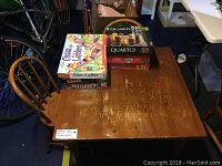 Photo showing worn wooden kids table with two matching wooden chairs and six board games stacked on the table including Quarto, Chutes and Ladders, Clue, and Battleship.
