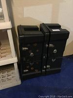 Two stacked large black plastic storage trunks with metal latches and no keys. One trunk decorated with craft paints and glued pieces.