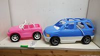 Two plastic toy vehicles, pink convertible and blue and white SUV, displayed on shelf.