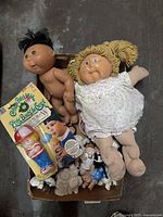 Top view photo showing two Cabbage Patch Kids dolls, vintage Fisher-Price boxed toy, ceramic figurines and various small accessories in a box.