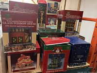 Boxes of porcelain lighted glass window houses with Christmas motif and a candlestick holder behind them. Some boxes show wear and tear.