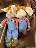 Photo of two plush dolls with striped red and white shirts, blue overalls, yellow yarn hair, large floppy ears, displayed side by side in cardboard box.