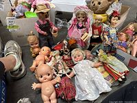 Overview photo of many vintage dolls including Kewpie dolls, ethnic fabric dolls, boxed items, and a stuffed bear. Shows condition and variety.