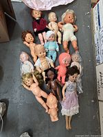Full set of 13 dolls laid out on a gray floor, showing different sizes, materials, and conditions, some with clothing, some nude or partially dressed.