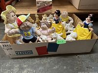 Box of assorted figurines including Disney Snow White characters, Wade, and bisque ceramics on a concrete floor.