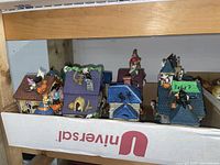 Overall group of 6 ceramic Halloween houses arranged in two rows inside a cardboard box showing various roof colors and Halloween decorations