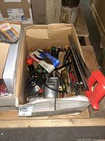 Box containing assorted hand tools including Speedway power drill, saws, pliers, wrenches, a hatchet, and cutting pliers in packaging. Some wear and rust visible.