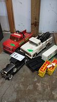 Photo showing red tow truck, white Buddy L tow truck with black crane, black vintage style car, white crates and other toy parts on floor